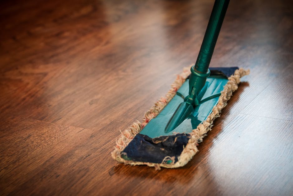 A light blue, teardrop-shaped wall hook mounted on a matte grey wall, holding a small wooden-handled brush with natural bristles. The brush is positioned vertically with the bristles facing downward, 