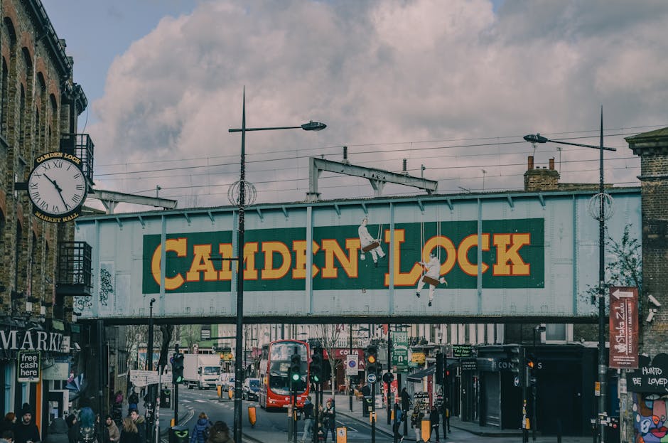 Exterior view of a multi-story brick building with large arched windows displaying the words 'CAMDEN LOCK'. Below, a blue sign with white text welcomes visitors to 'CAMDEN LOCK'. The building houses s