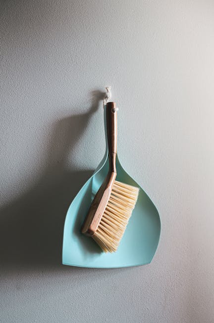A close-up of a muted beige wall with a small, minimalist corner shelf composed of a pastel blue, curved tray. Resting on the tray is a wooden-handled scrub brush with light bristles. The shelf appear